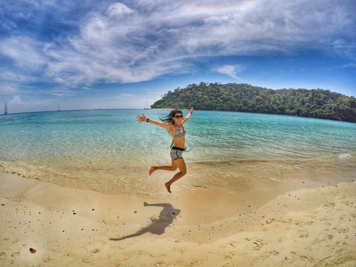 Person jumping joyfully on a sandy beach with turquoise water and a lush, green island in the background, in Thailand.