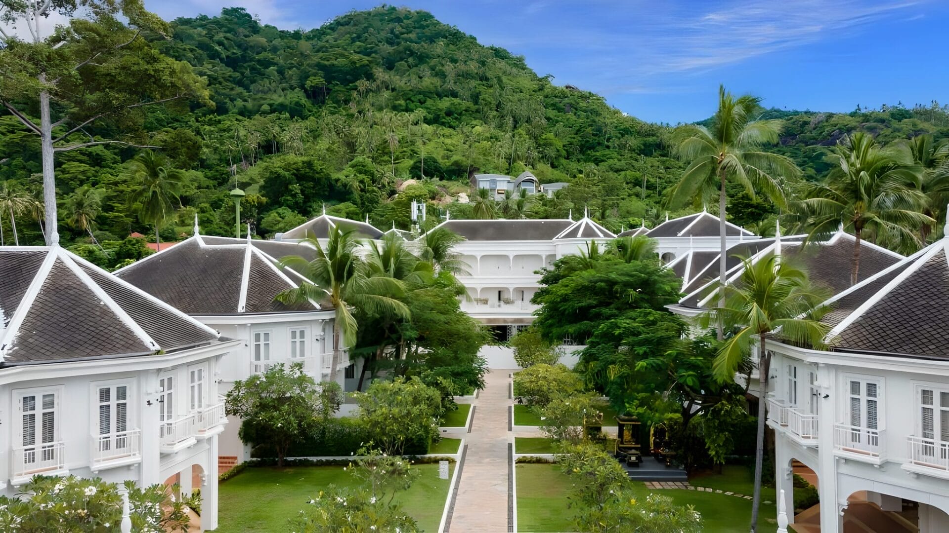 Aerial view of the Outrigger Koh Samui Beach Resort on Koh Samui, Thailand, surrounded by lush green hills and encircled by tall palm trees on a clear day.