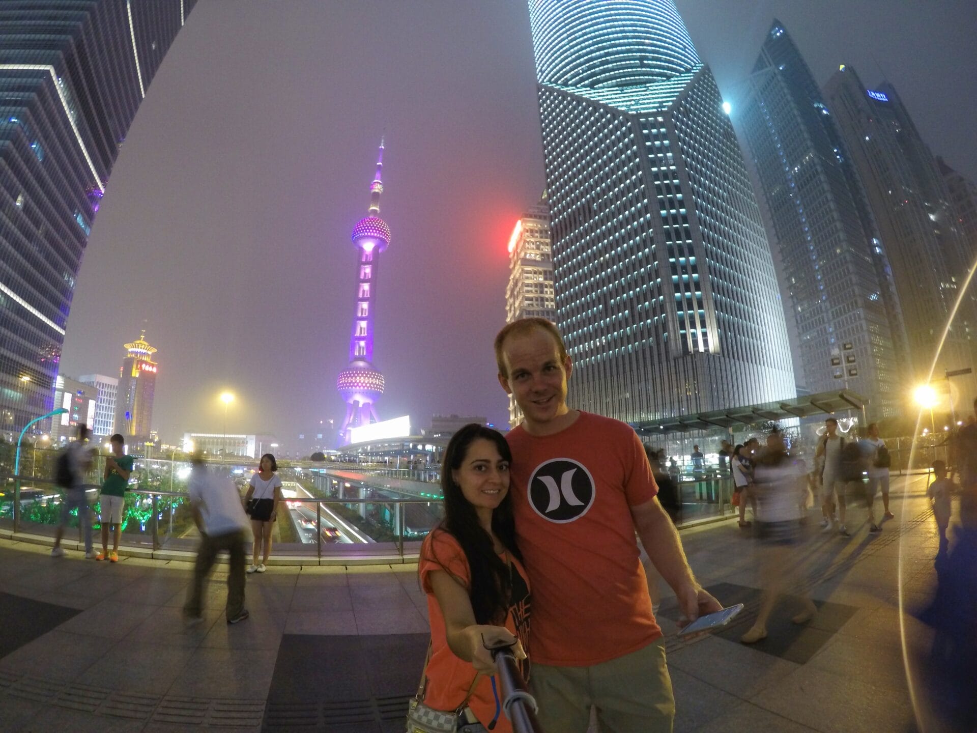 A couple taking a selfie in front of the illuminated skyline of Shanghai, China, with the Oriental Pearl Tower prominently visible in the background.